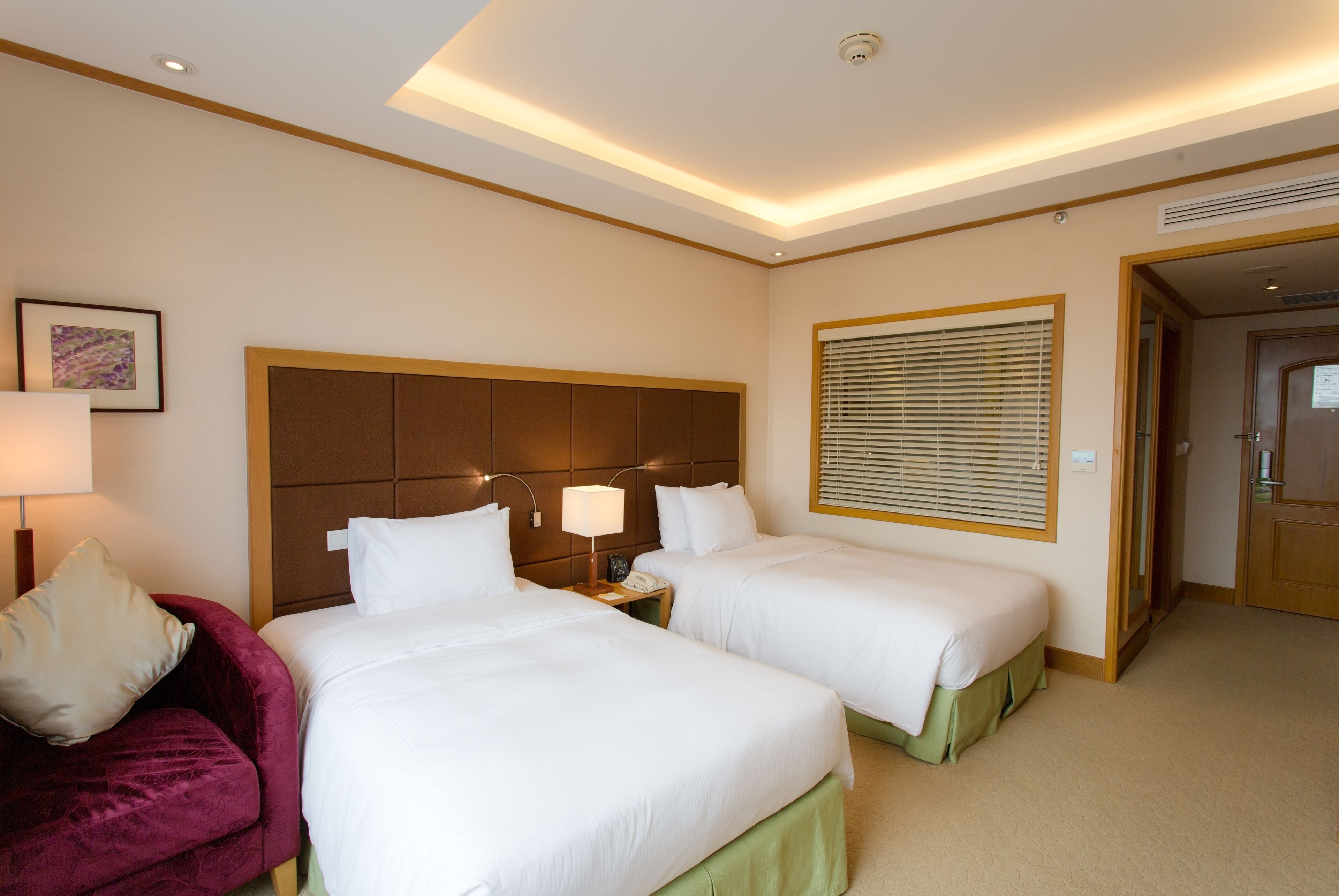 A hotel room with two beds, a red sofa, a bedside table with a lamp, and a window with blinds.