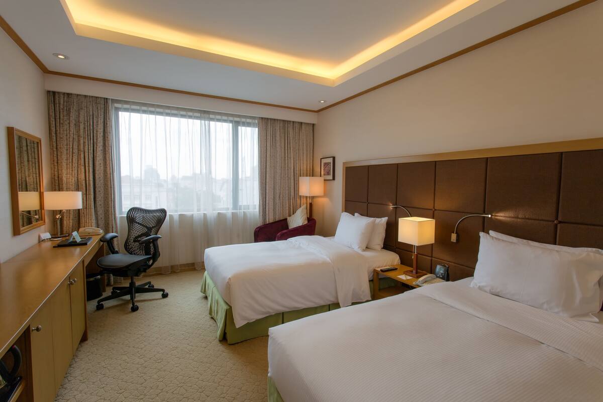 A hotel room with two beds, a desk, a chair, and a large window with curtains.