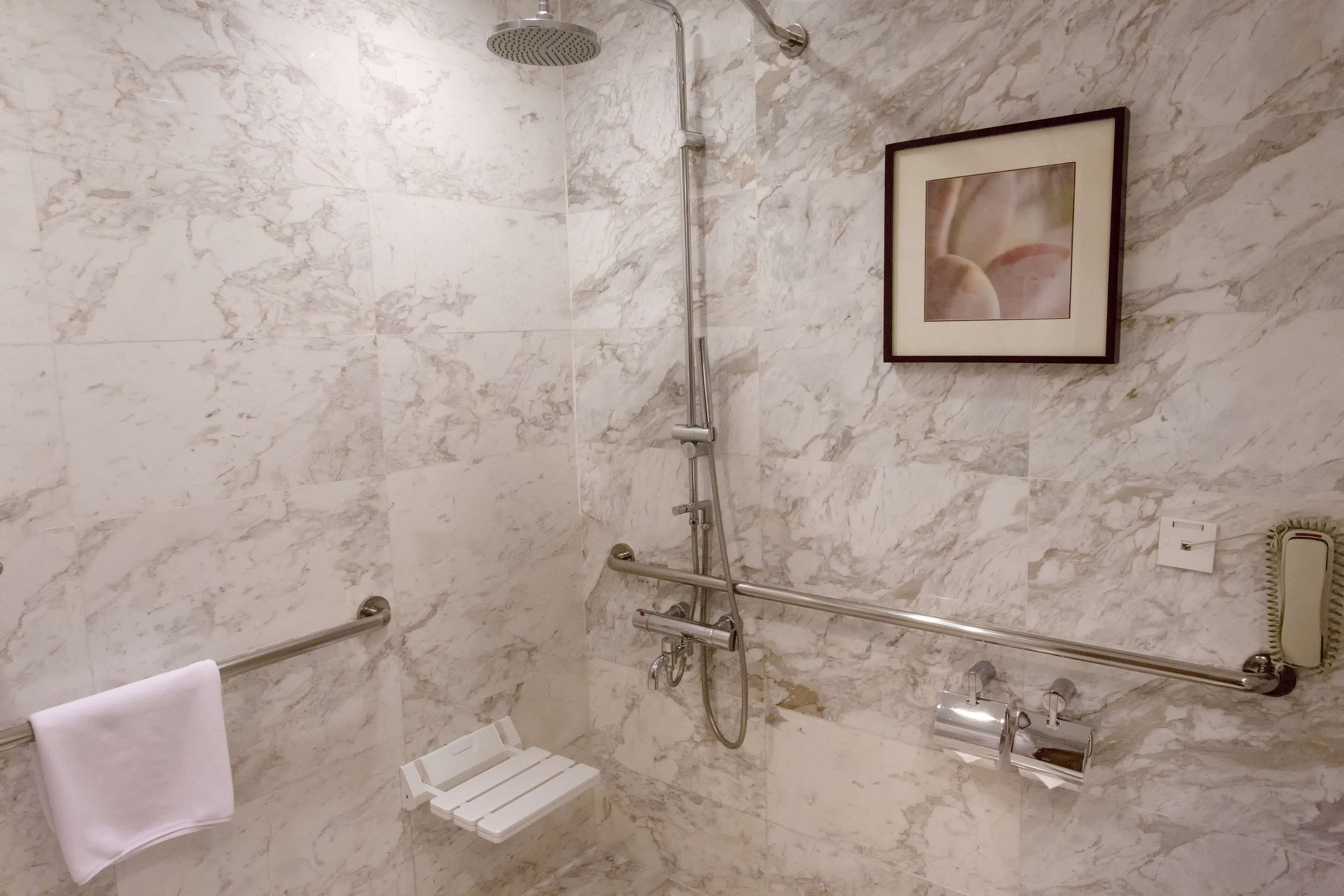 A shower area with a handheld showerhead, a glass-enclosed shower, a towel rack, a framed picture on the wall, and a small white bench.