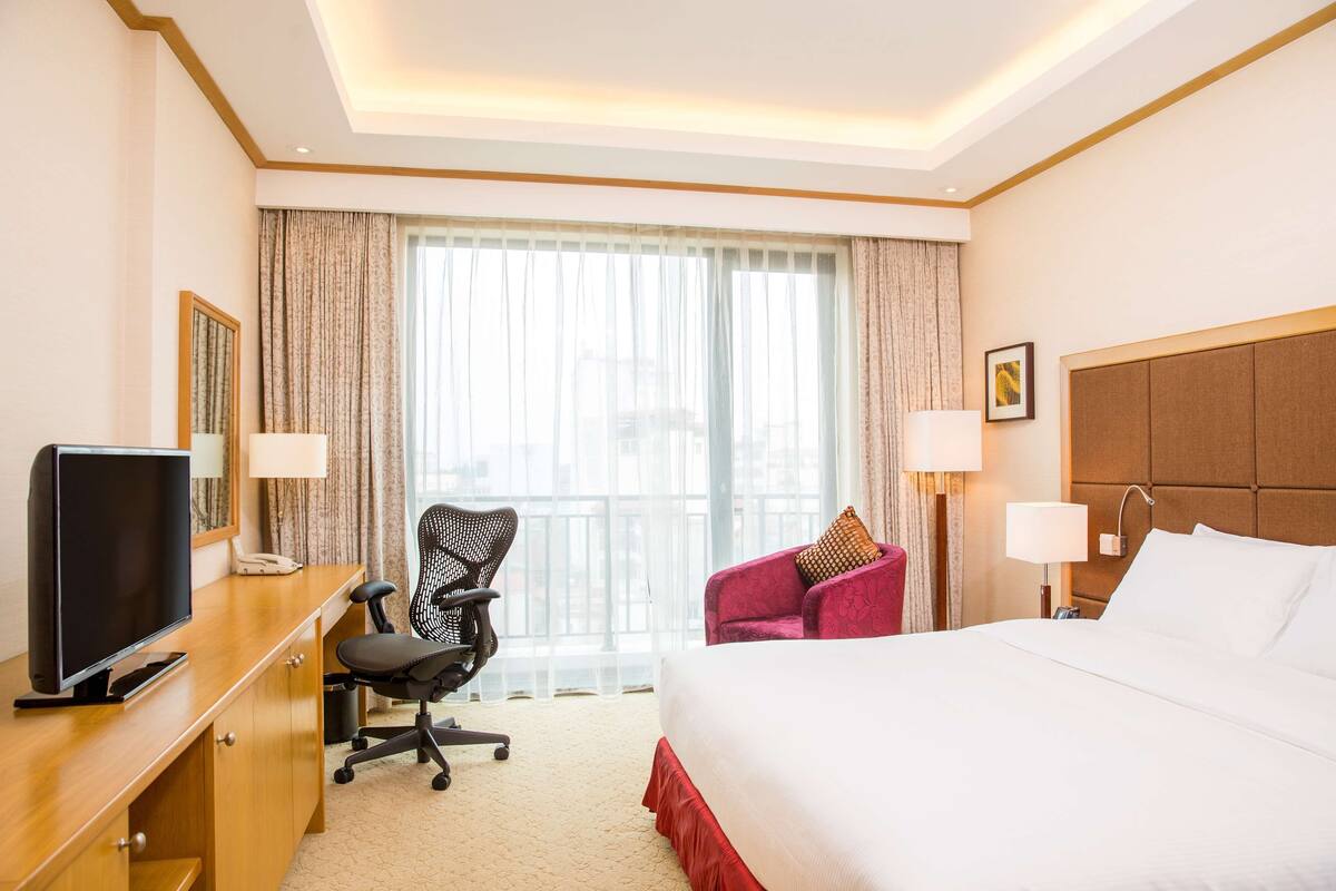 A hotel room with a bed, a desk with a TV, a chair, and a window with curtains.