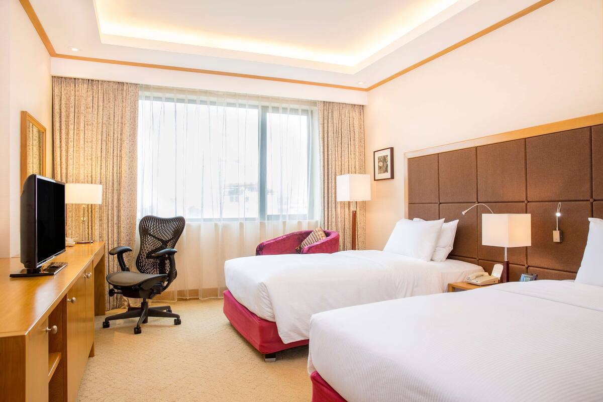 A hotel room with two beds, a desk with a TV, a chair, and a large window with curtains.
