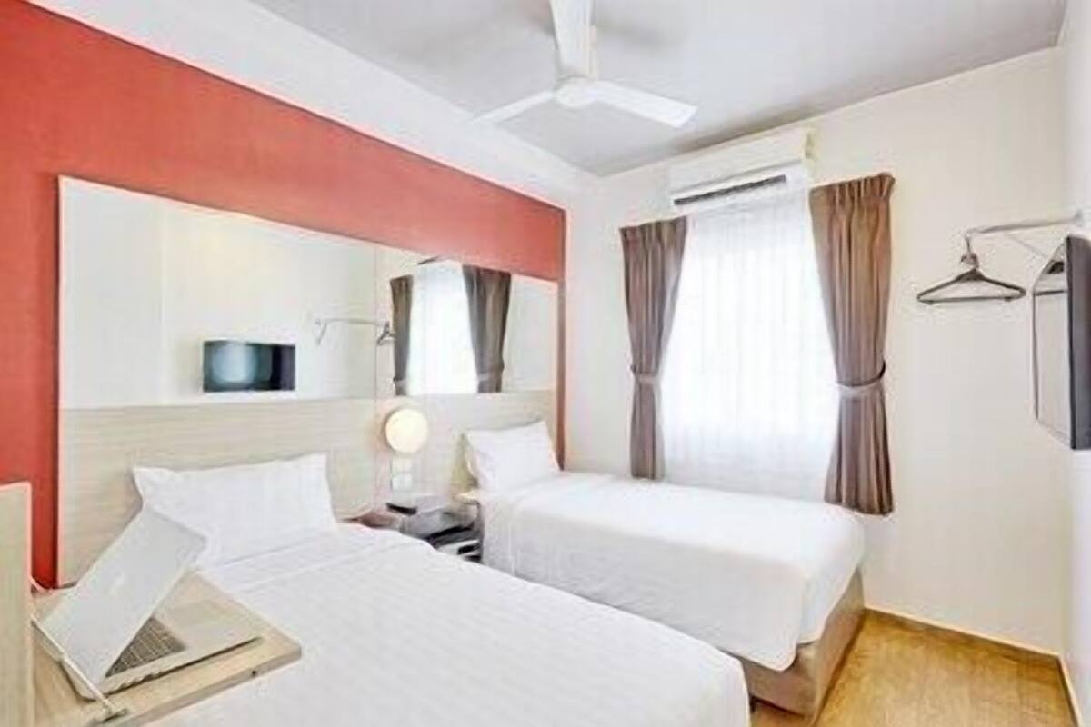 A hotel room with two beds, a desk with a laptop, a mirror, a TV, and an air conditioning unit.
