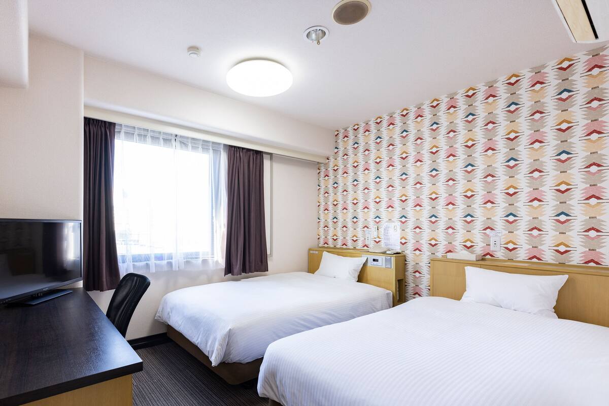 A hotel room with two beds, a desk, a TV, and a patterned wall.