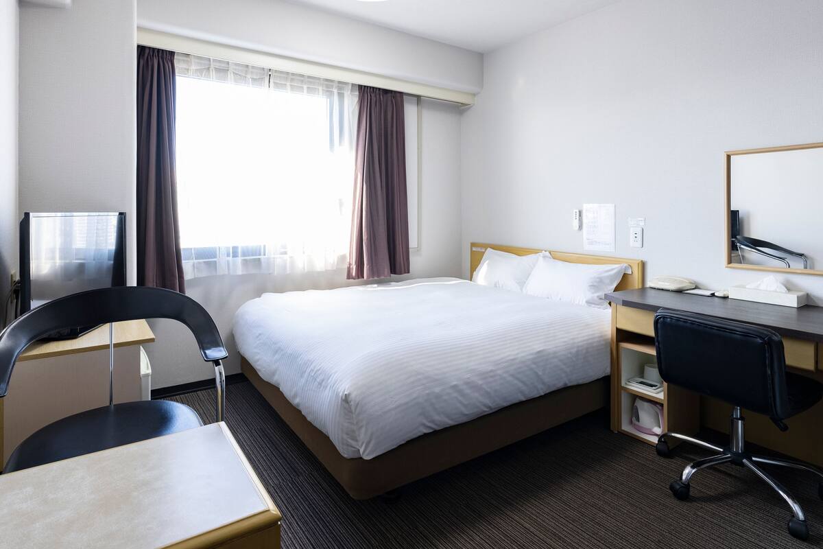 A hotel room with a bed, a desk, a chair, a TV, and a window with curtains.