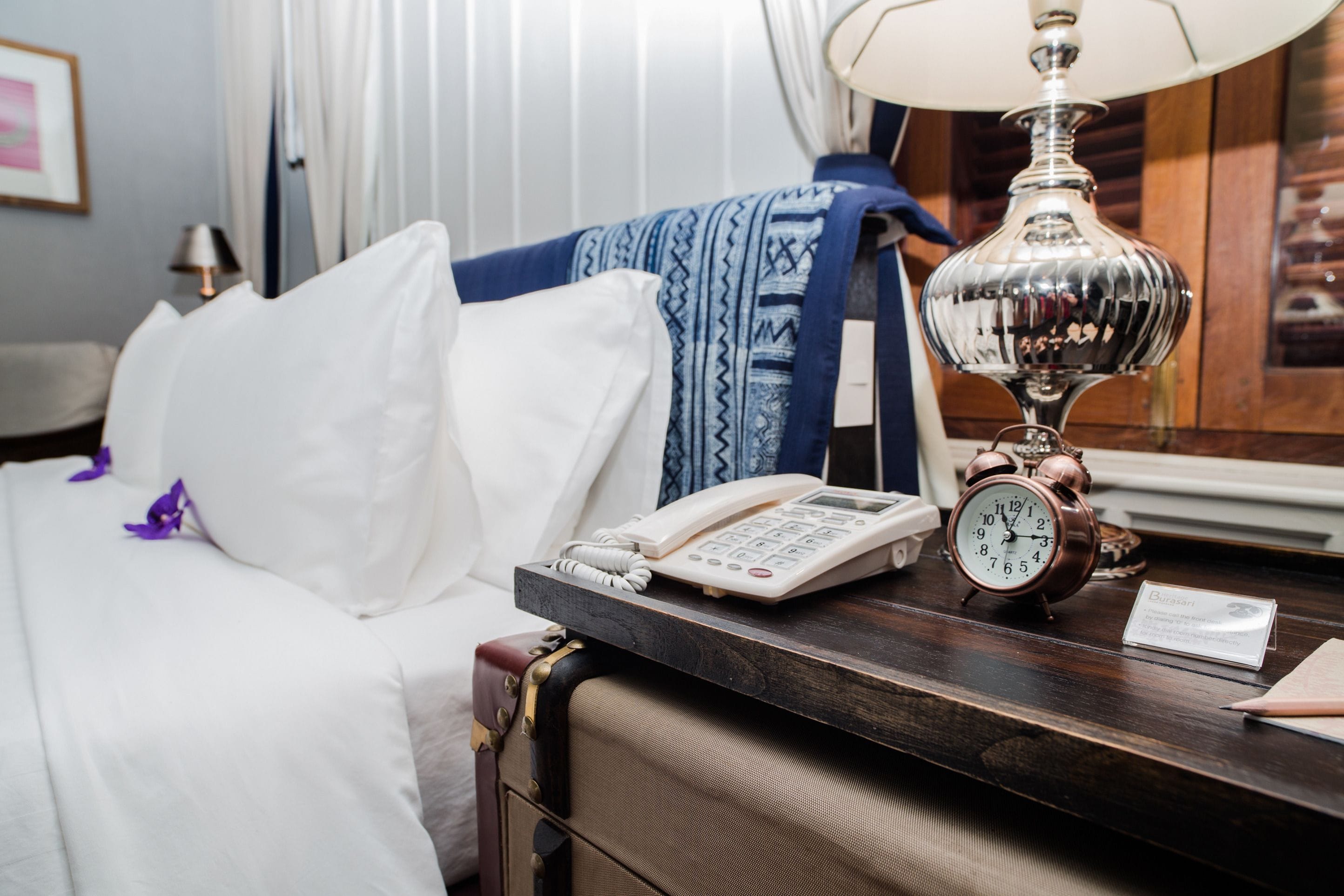 A hotel room with a bed, a bedside table with a lamp, a telephone, and a clock.