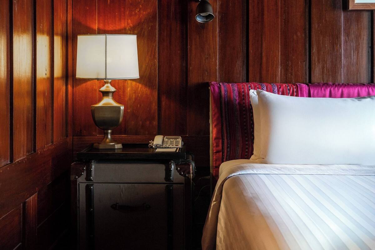 Habitación de hotel con pared de paneles de madera, una lámpara de noche, un teléfono y una cama con cubrecama blanco y manta a rayas rosadas y rojas.