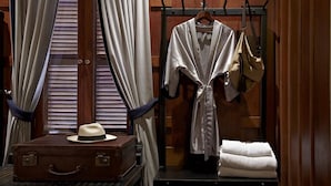 A room with a wooden wardrobe, a hanging coat rack with a robe and bag, a suitcase with a hat, and a window with blinds. - Burasari Heritage Luang Prabang (Luang Prabang)