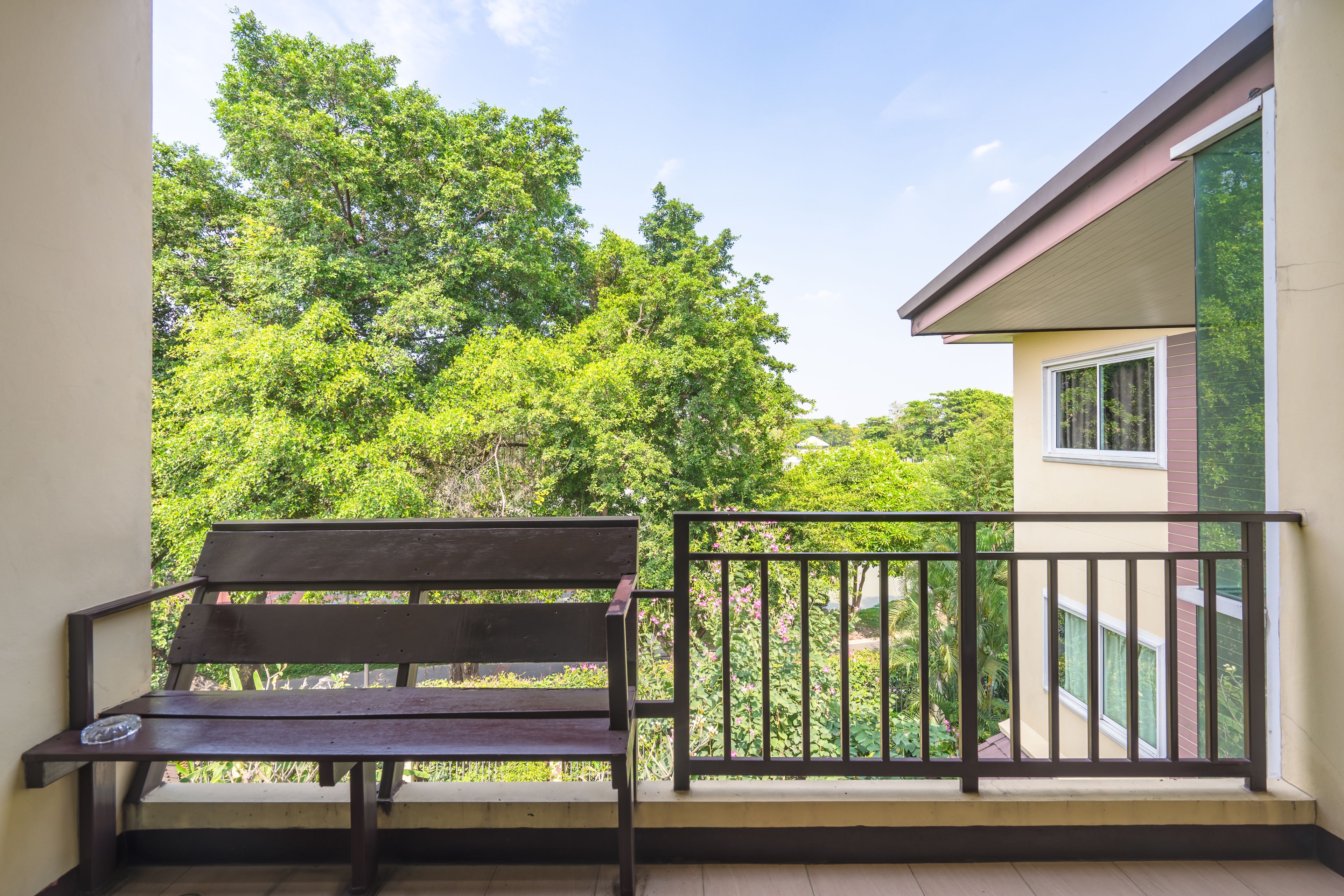 A balcony with a wooden bench and a black metal railing, overlooking a lush green area with trees and a building in the background.