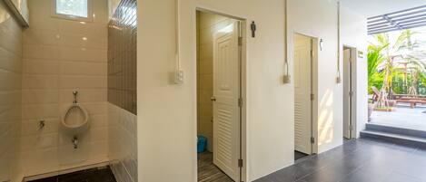 Ein modernes Badezimmer mit Pissoir, Dusche und Toilette. Eine Tür führt zu einem Balkon mit Blick auf Palmen.