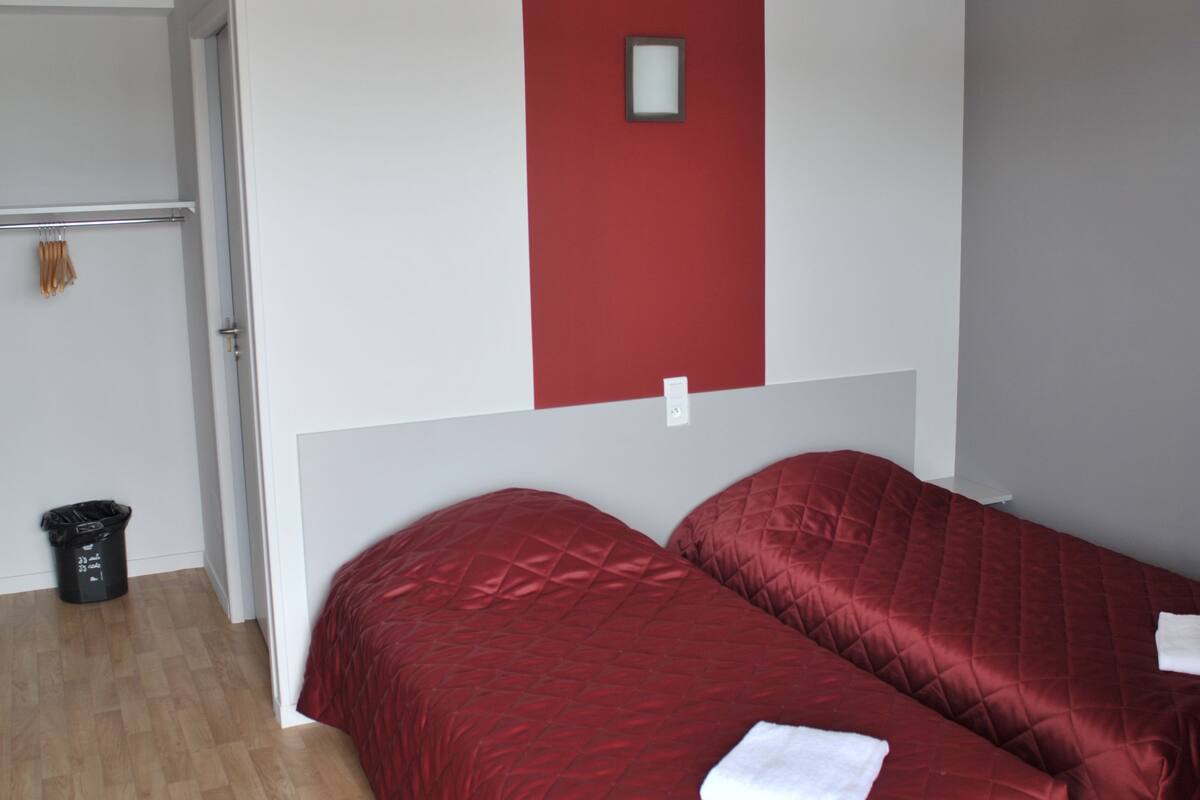 A room with two beds, a red accent wall, a white ceiling, and a wooden floor.