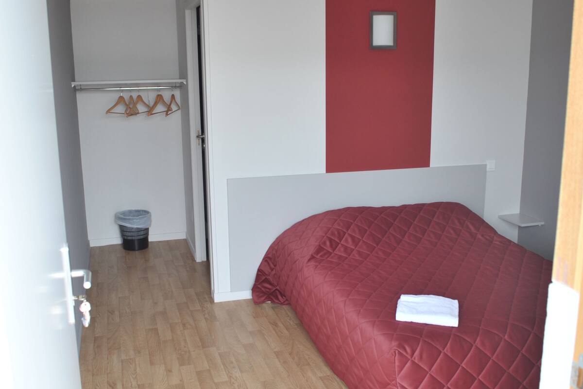 A bedroom with a red bedspread, a white wall with a red accent, a wooden floor, a small trash bin, and a coat rack with hangers.
