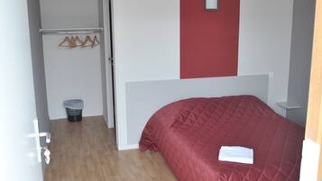 A bedroom with a red bedspread, a white wall with a red accent, a wooden floor, a small trash bin, and a coat rack with hangers.