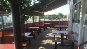 An outdoor patio area with wooden tables and benches, surrounded by glass walls and a view of the street.