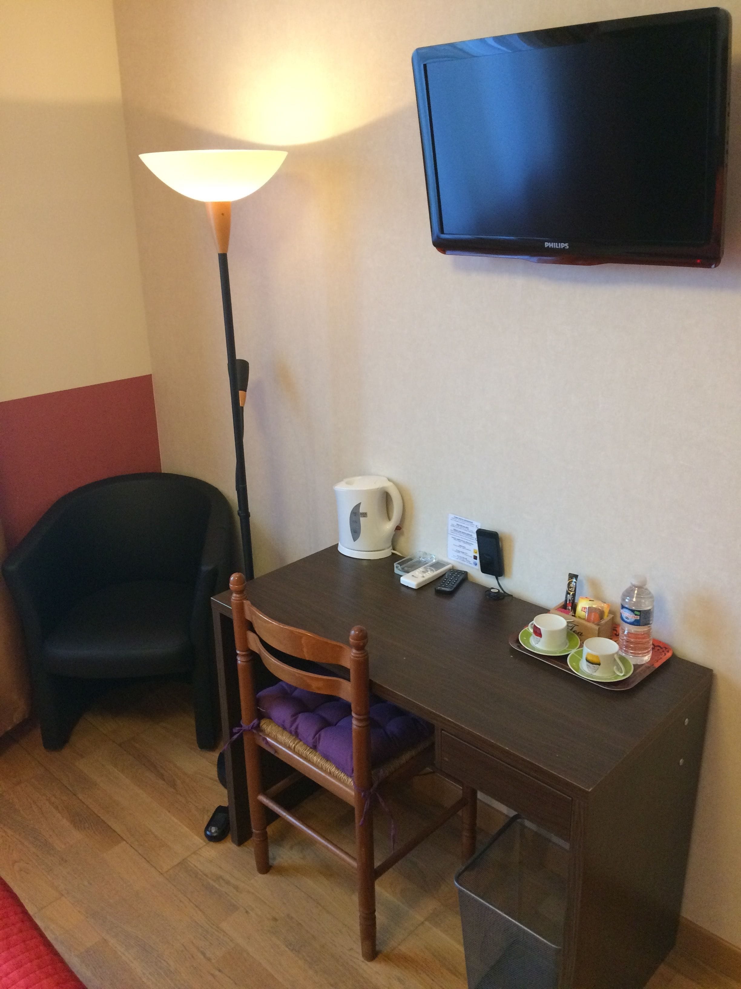 Une petite pièce avec un bureau en bois, une chaise, une lampe, un téléviseur et un plateau contenant des tasses et une bouteille.