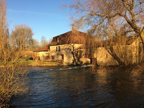 A rustic house by a river with a wooden deck and a car parked nearby.