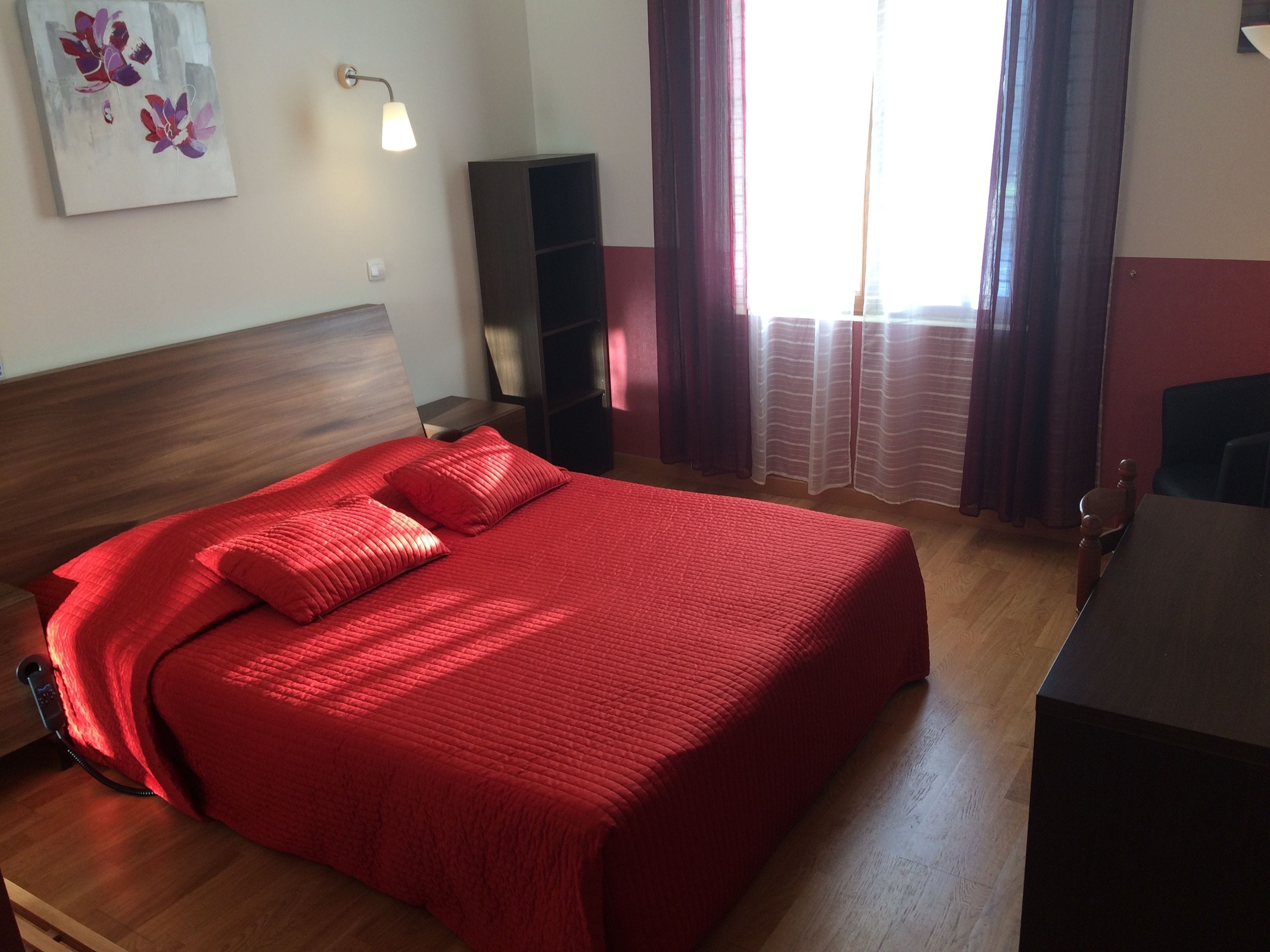 A neatly made bed with red bedding, a bedside table, a lamp, a bookshelf, and a window with curtains.
