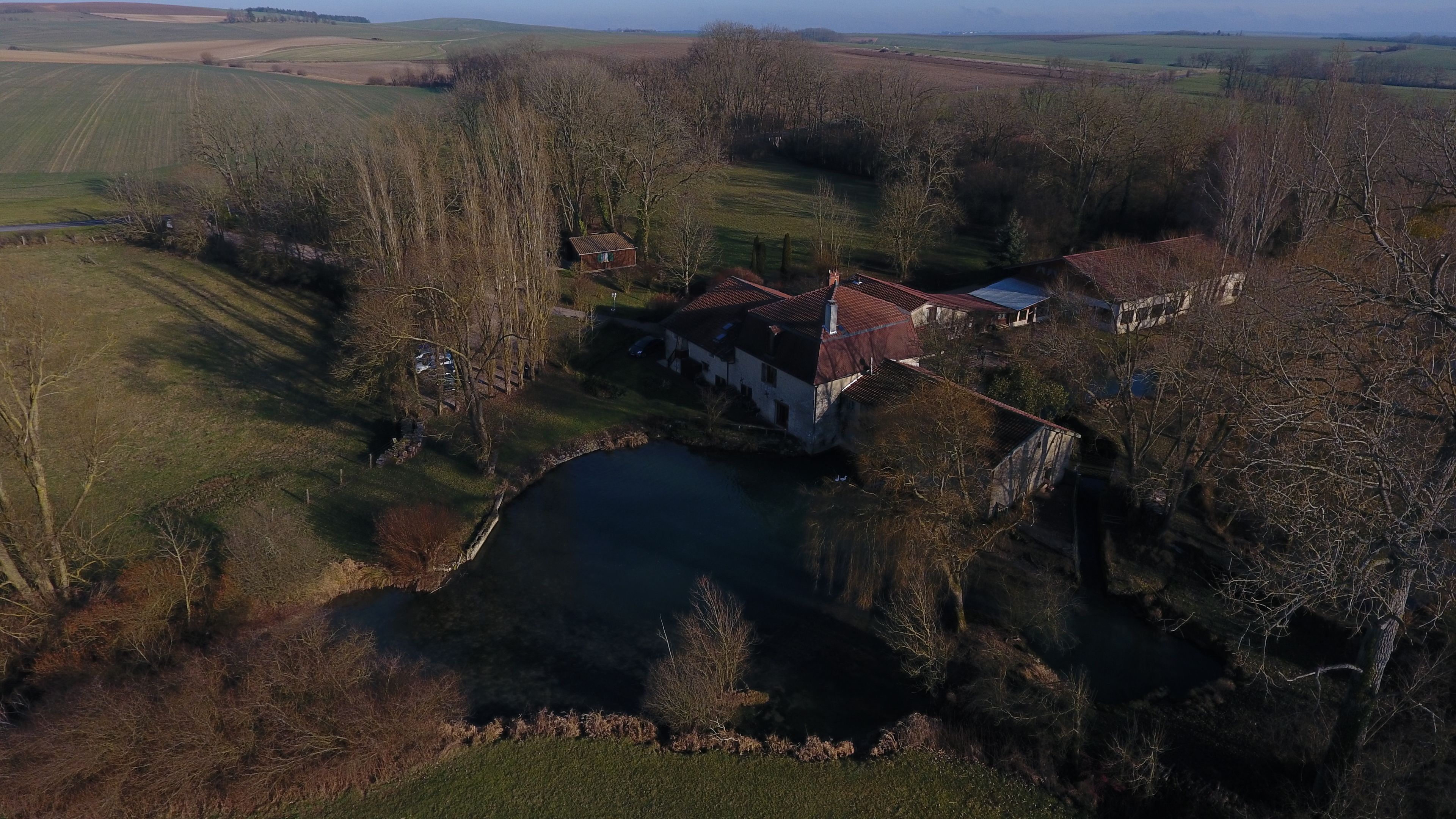 Luchtbeeld van een landgoed met een groot vijver, een huis met een rood dak en omliggende velden.