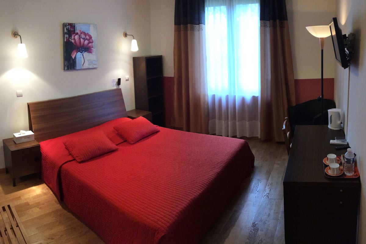 Habitación de hotel con una cama grande, una mesita de noche, un escritorio con lámpara, un cuadro en la pared y una ventana con cortinas.