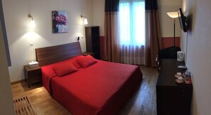 A hotel room with a large bed, a nightstand, a desk with a lamp, a painting on the wall, and a window with curtains.