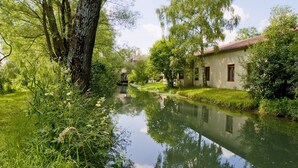 Un paisaje tranquilo a orillas del río con un edificio tradicional de teja roja, rodeado de abundante vegetación y una corriente de agua en calma que refleja los árboles.