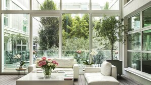 Un salon moderne avec un canapé blanc, une table basse agrémentée de livres et de fleurs, et de grandes fenêtres donnant sur un paysage verdoyant et un balcon.