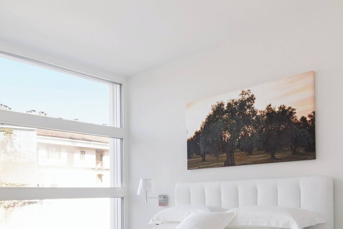 A modern bedroom with a large bed, a nightstand, a wall-mounted lamp, and a window with a view of buildings.