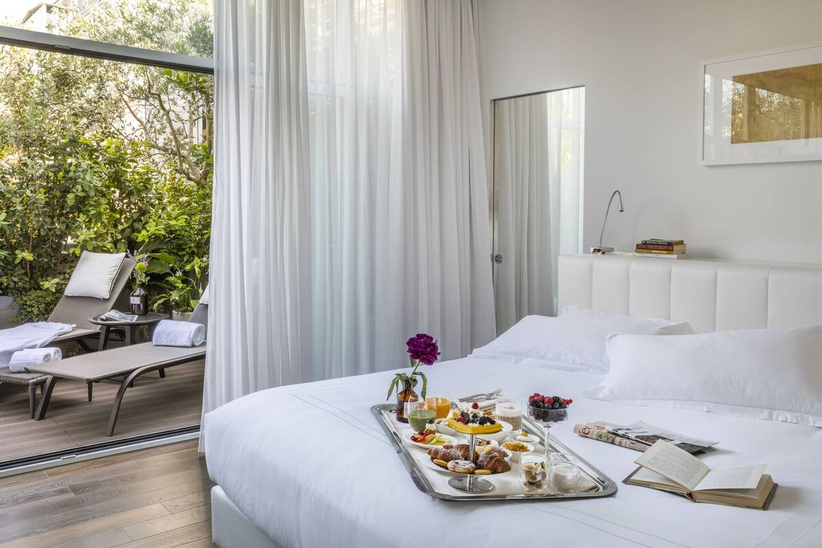 A hotel room with a bed, a tray of breakfast items, a window with curtains, and a view of greenery outside.