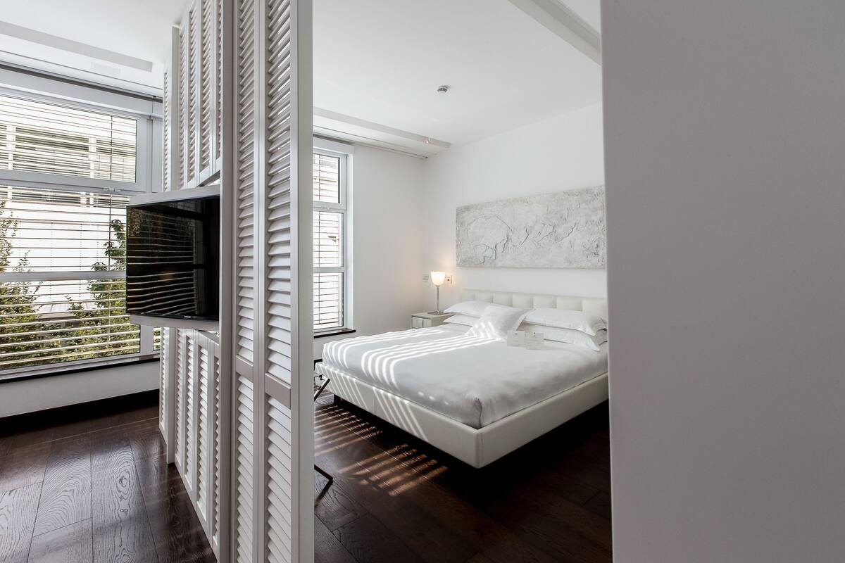 A bedroom with a large bed, a nightstand, a wall-mounted light, and a window with blinds.