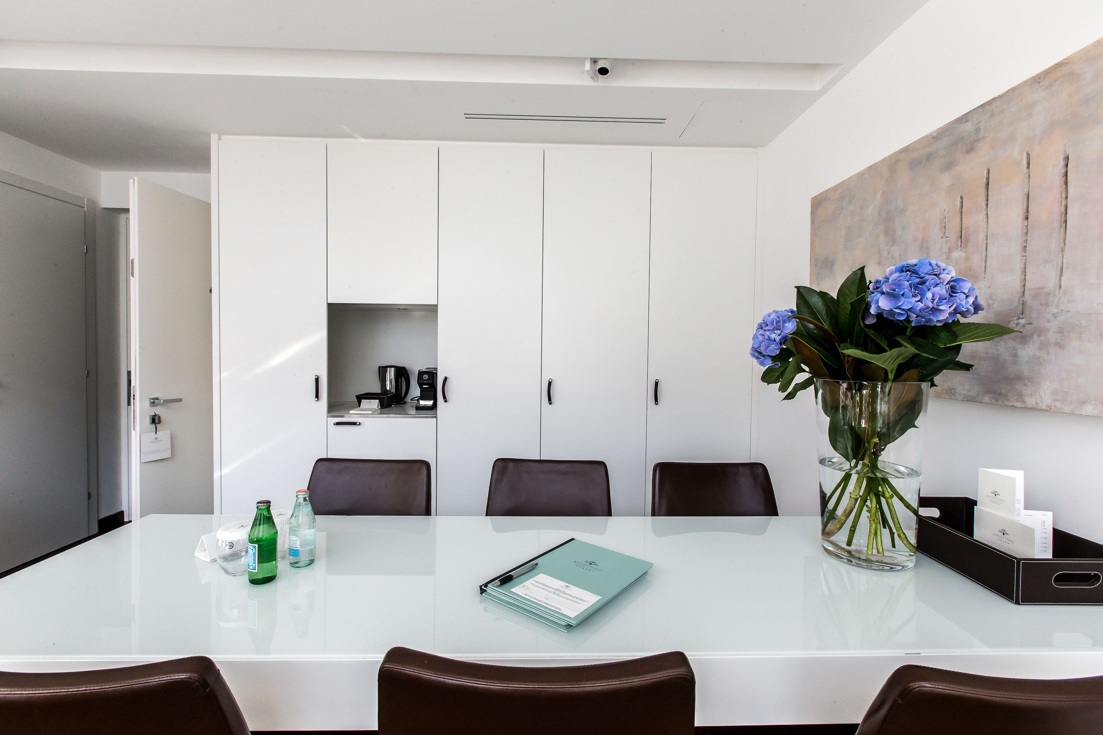 Una sala de conferencias moderna con una mesa blanca, sillas marrones, un florero con flores y un cuadro en la pared.