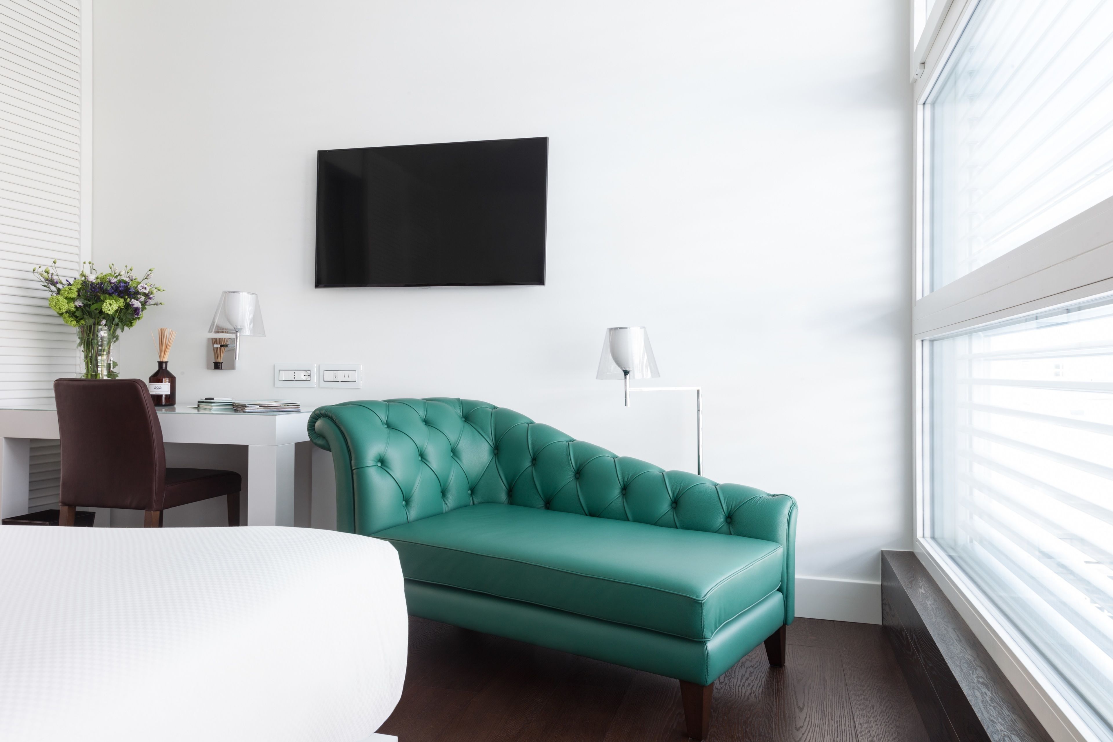 A modern hotel room with a teal sofa, a flat-screen TV, a desk with a chair, and a large window with blinds.