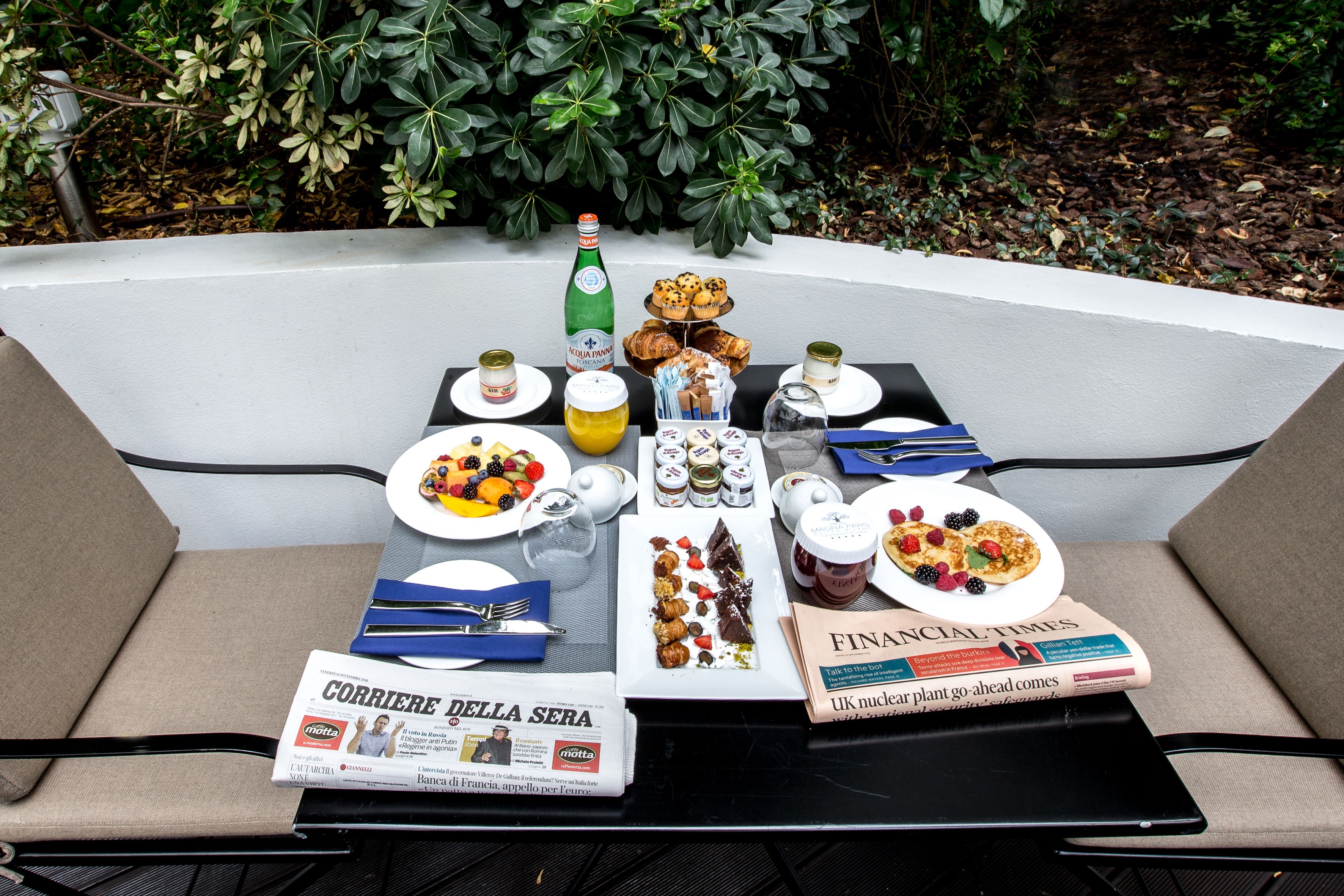 Une table à manger installée à l’extérieur, avec des assiettes remplies de nourriture, une bouteille et des journaux.