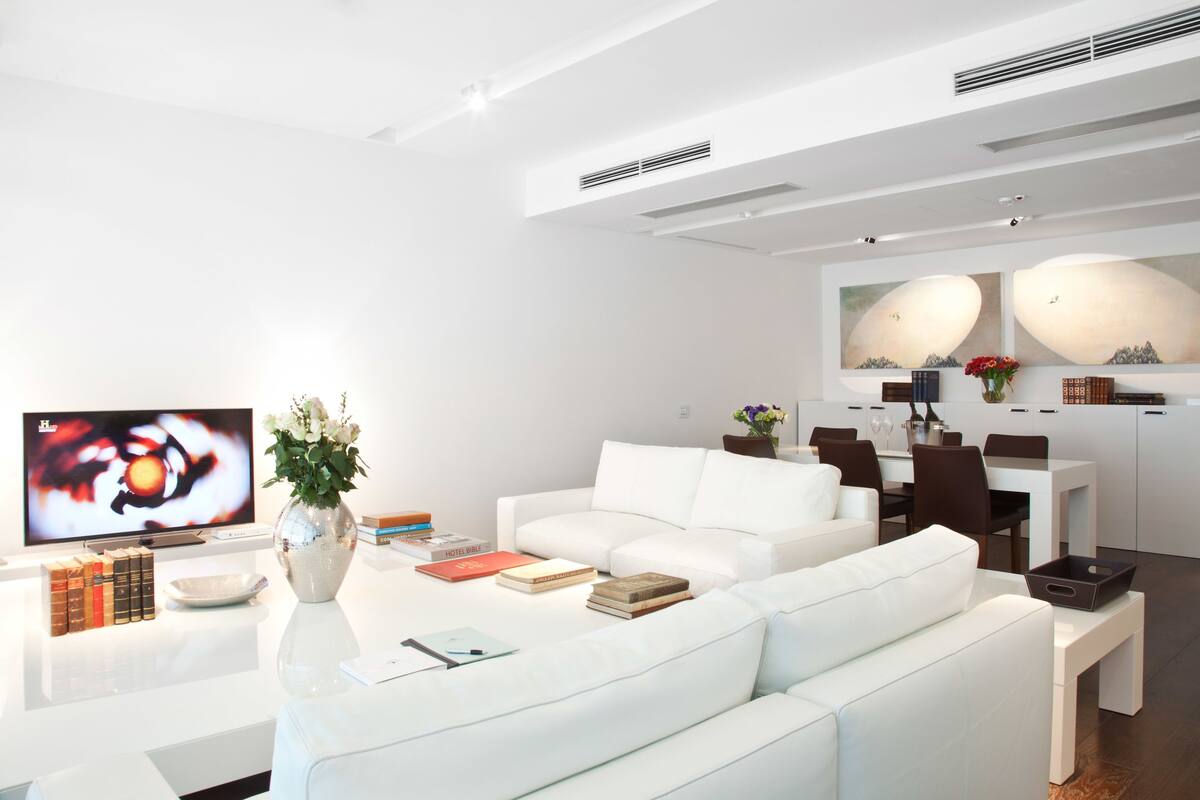 A modern living room with a white sofa, a flat-screen TV, a vase with flowers, and a dining area with a table and chairs.