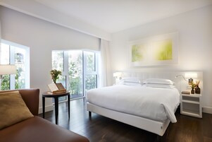 Una camera d'hotel moderna con un letto grande, una poltrona, un tavolino con un libro e fiori, e un comodino con una lampada e oggetti decorativi.