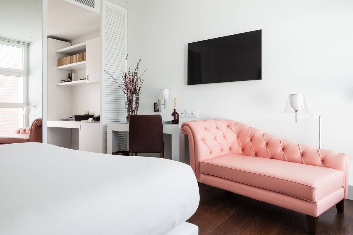 A modern bedroom with a large bed, a pink sofa, a desk with a chair, a TV on the wall, and a window with blinds.