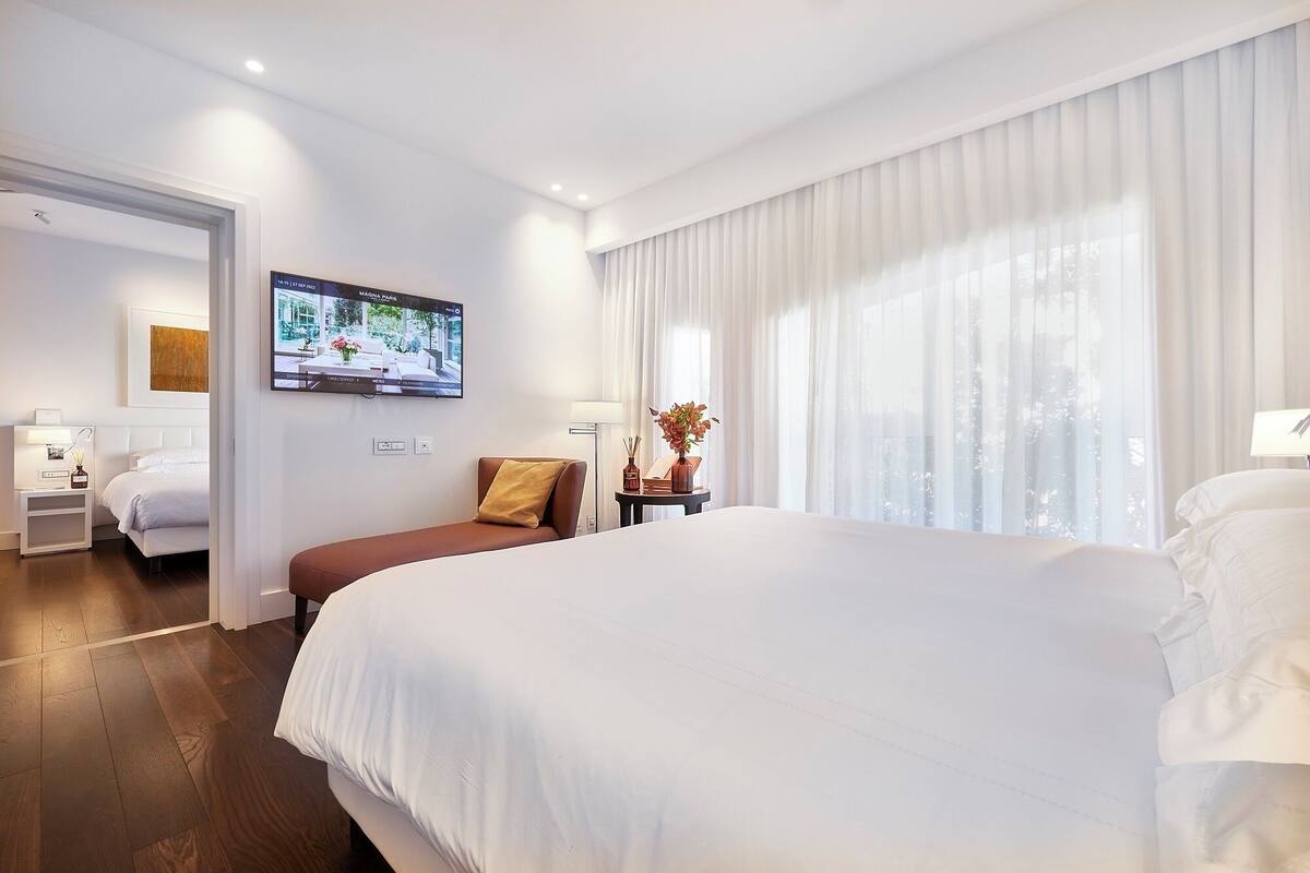 A hotel room with a large bed, a TV on the wall, a desk with a lamp, and a window with curtains.