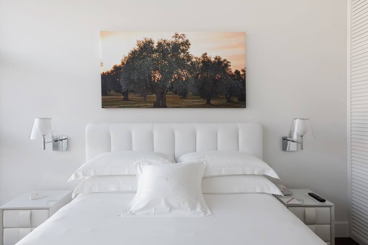 A neatly arranged bedroom with a large bed, white bedding, two bedside tables with lamps, and a wall-mounted painting of trees.