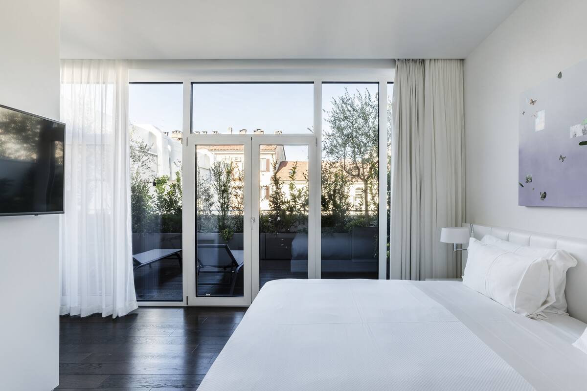 A modern hotel room with a large bed, a flat-screen TV, a sliding glass door to an outdoor area, and a view of the cityscape.