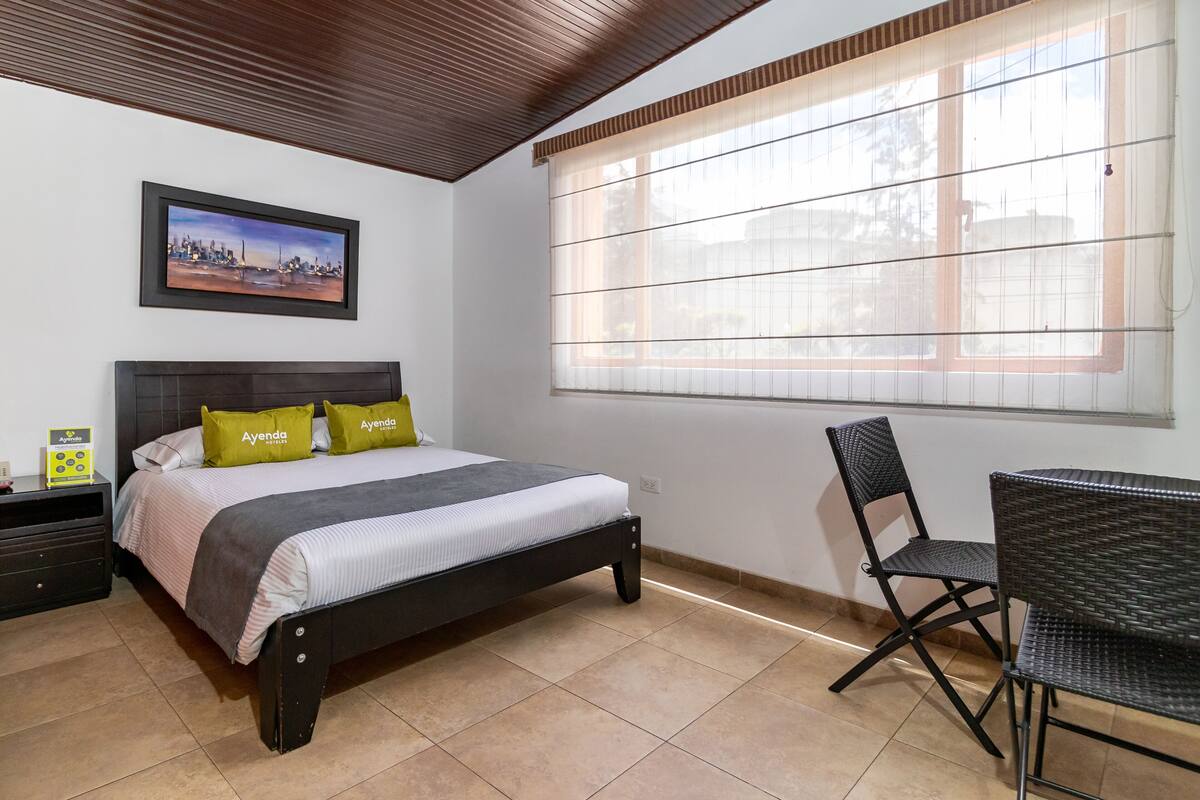 Un dormitorio con cama, una silla, una mesita de noche, una ventana con persianas y un cuadro en la pared.