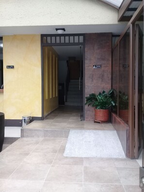 Ingresso di un hotel con pavimento piastrellato, tappetino di benvenuto, una pianta in vaso e una scala che conduce ai piani superiori.