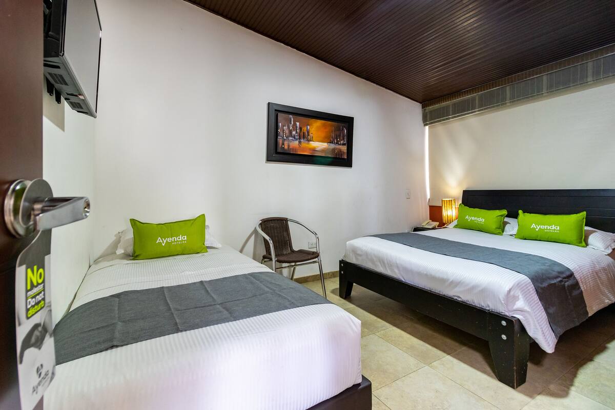 Habitación de hotel con dos camas, cada una con un almohadón verde con la etiqueta 'Ayenda'.