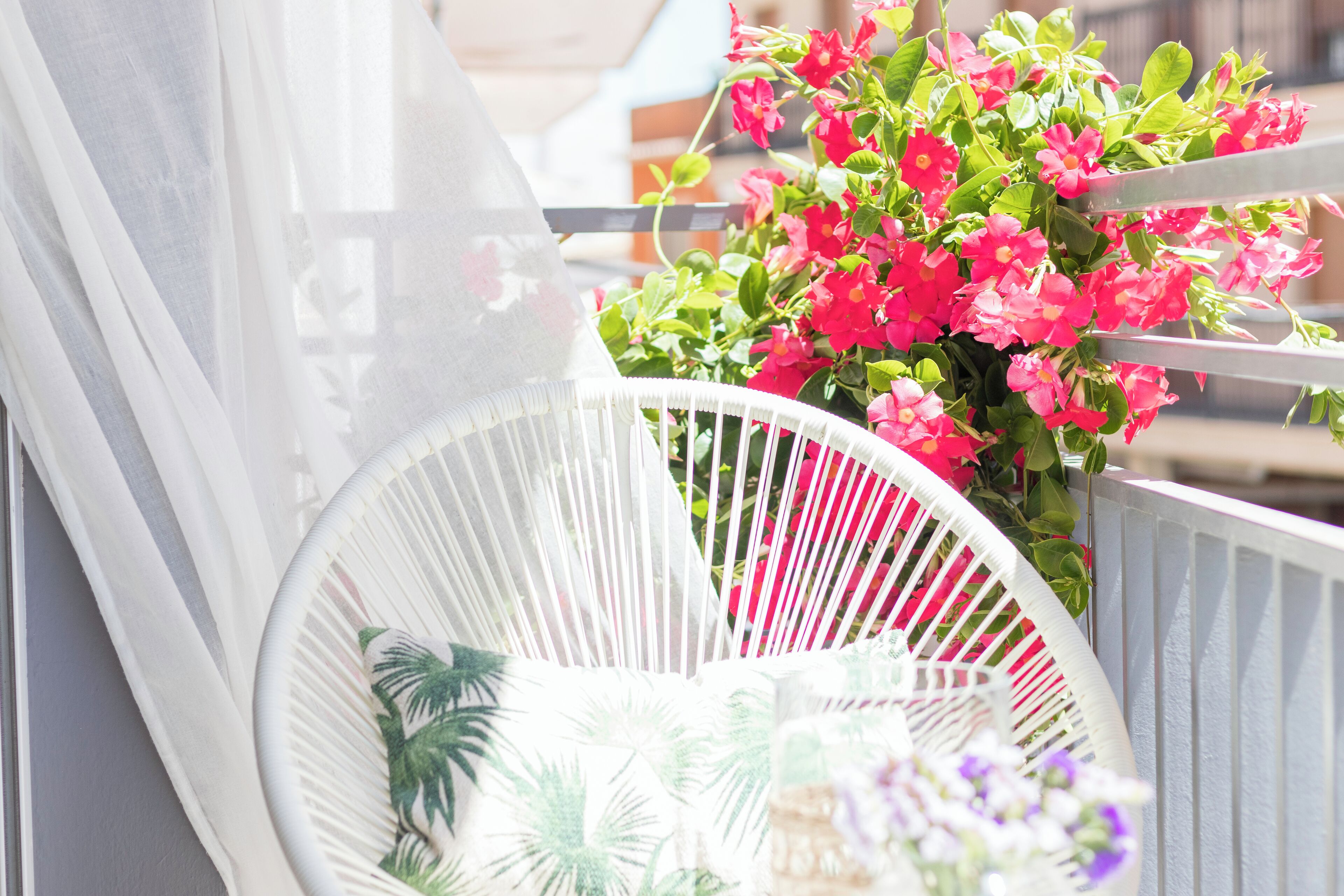 Una silla blanca de exterior con un cojín estampado tropical, ubicada junto a una baranda blanca adornada con flores rosadas.