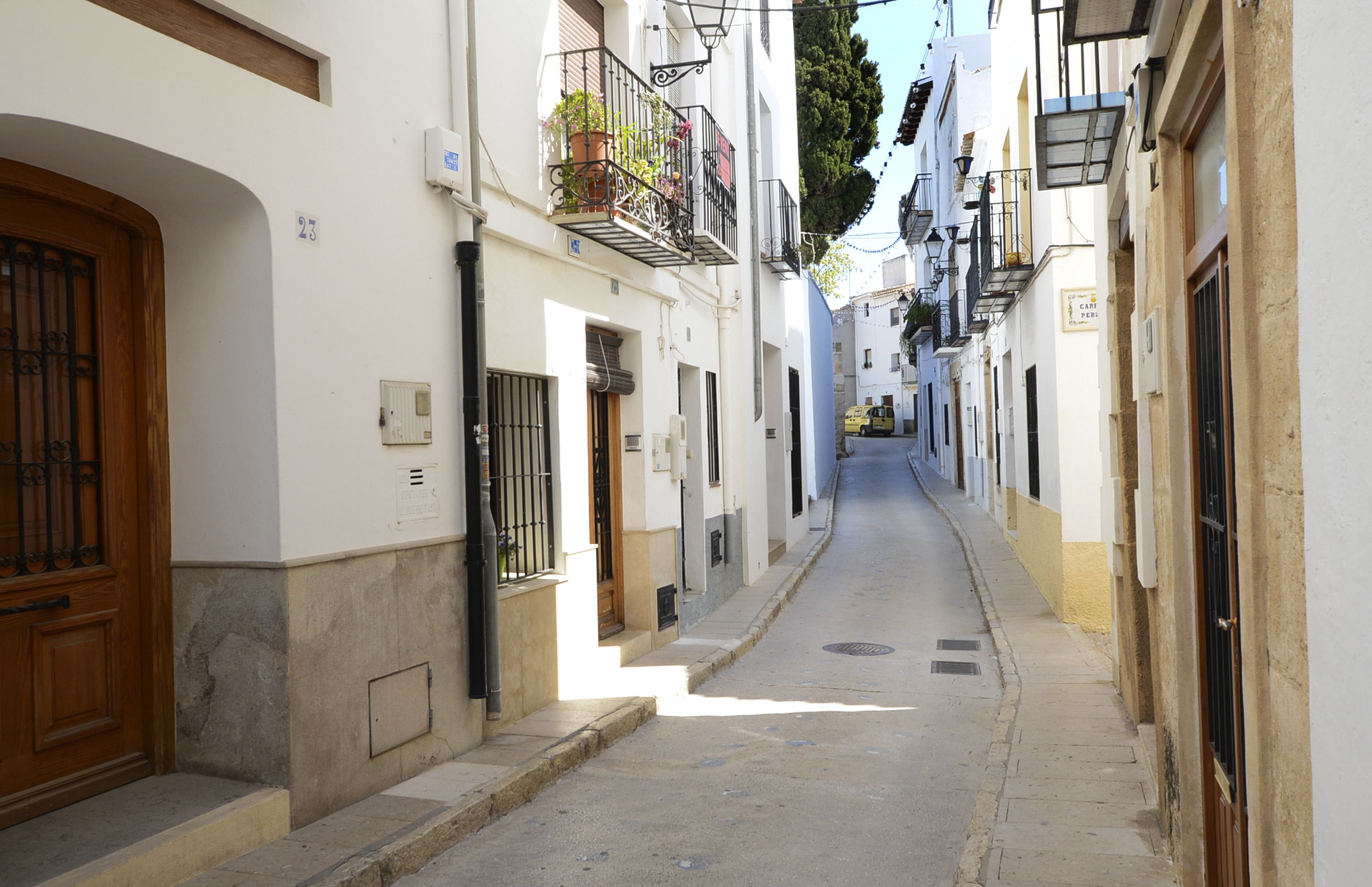 Una calle estrecha con edificios blancos, puertas de madera y balcones con plantas.