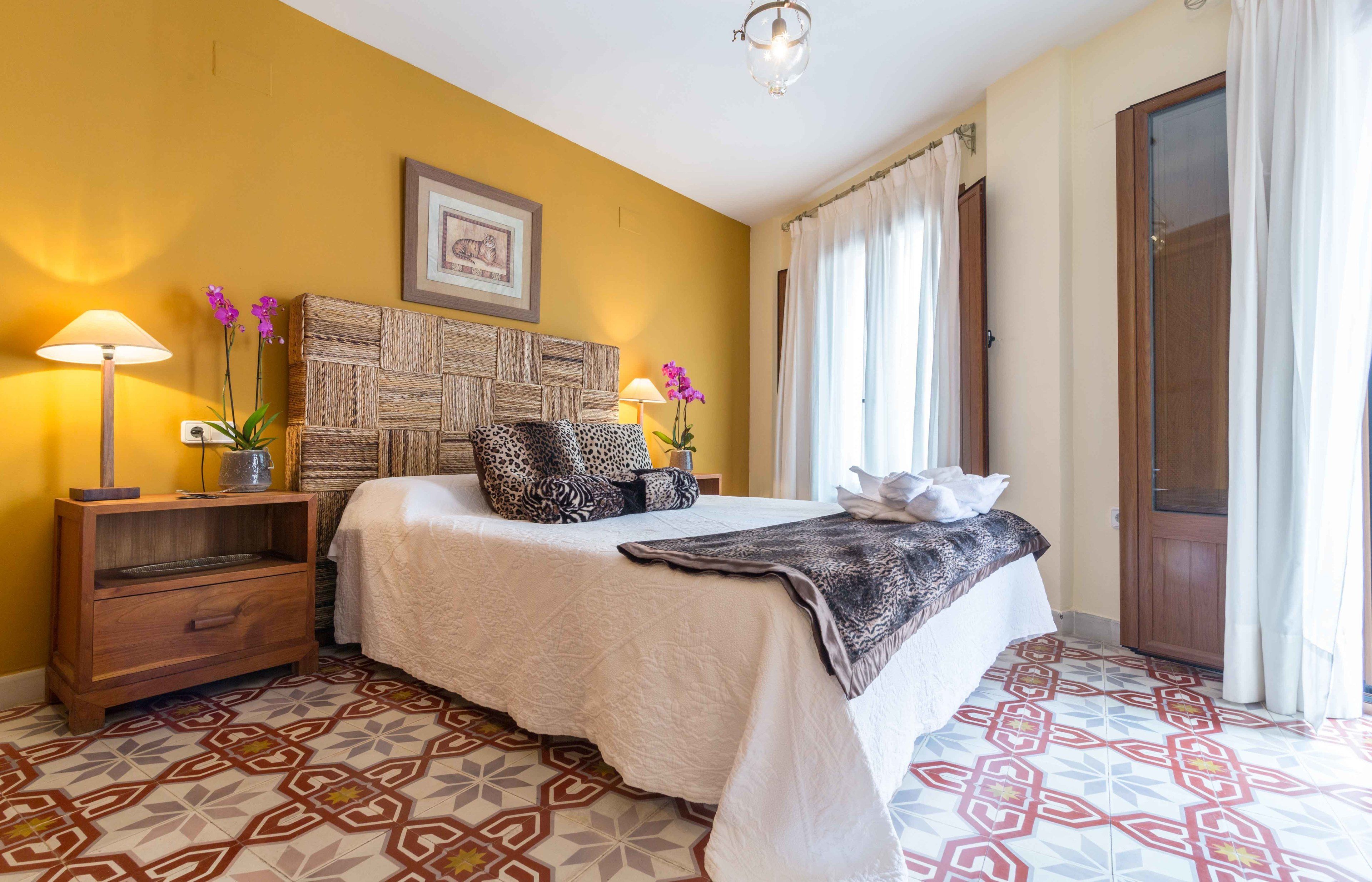 Una habitación de hotel con cama, mesita de noche, lámpara, un jarrón con flores y piso estampado.