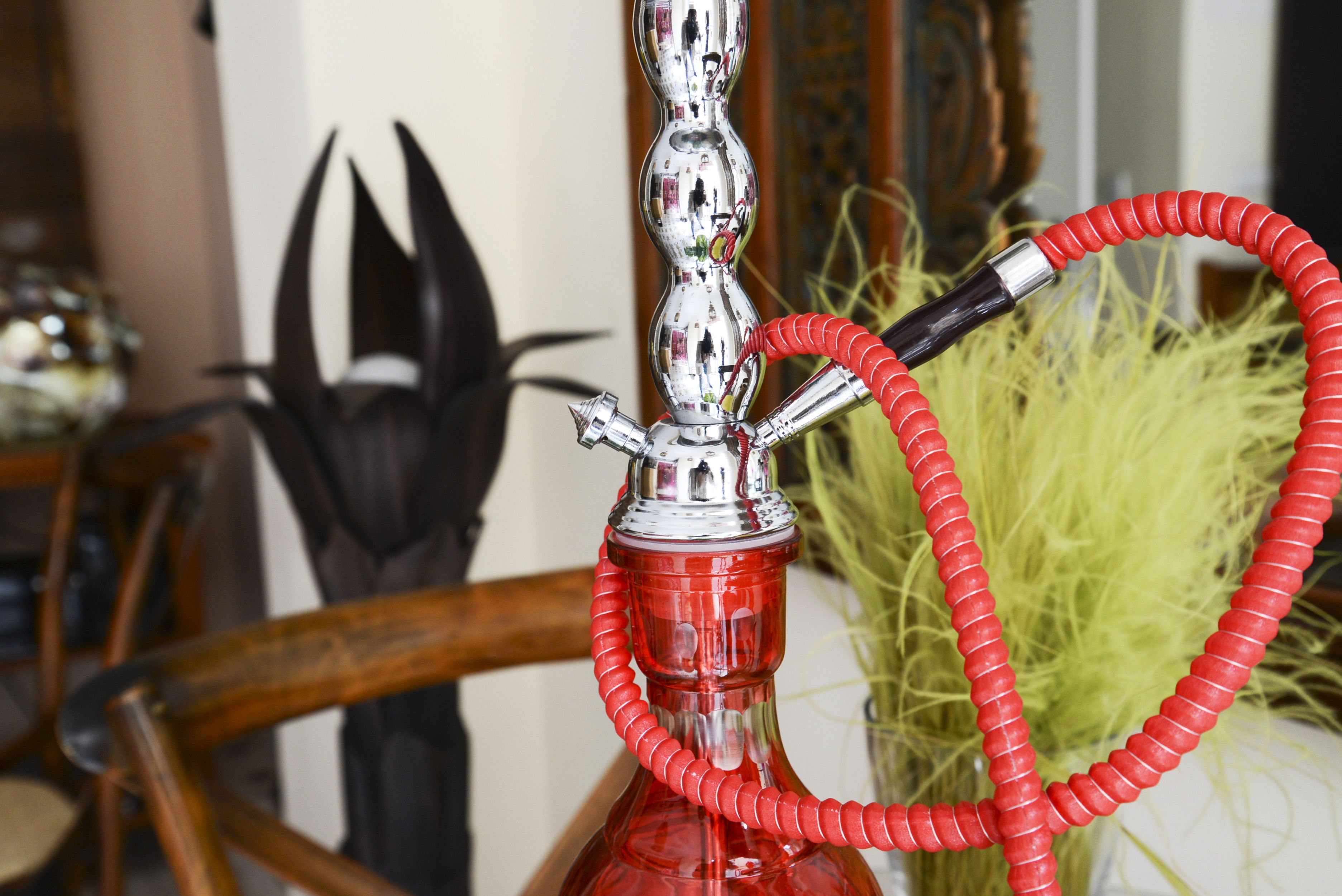 A hookah with a red hose and a metallic base, placed on a wooden surface with a decorative item in the background.