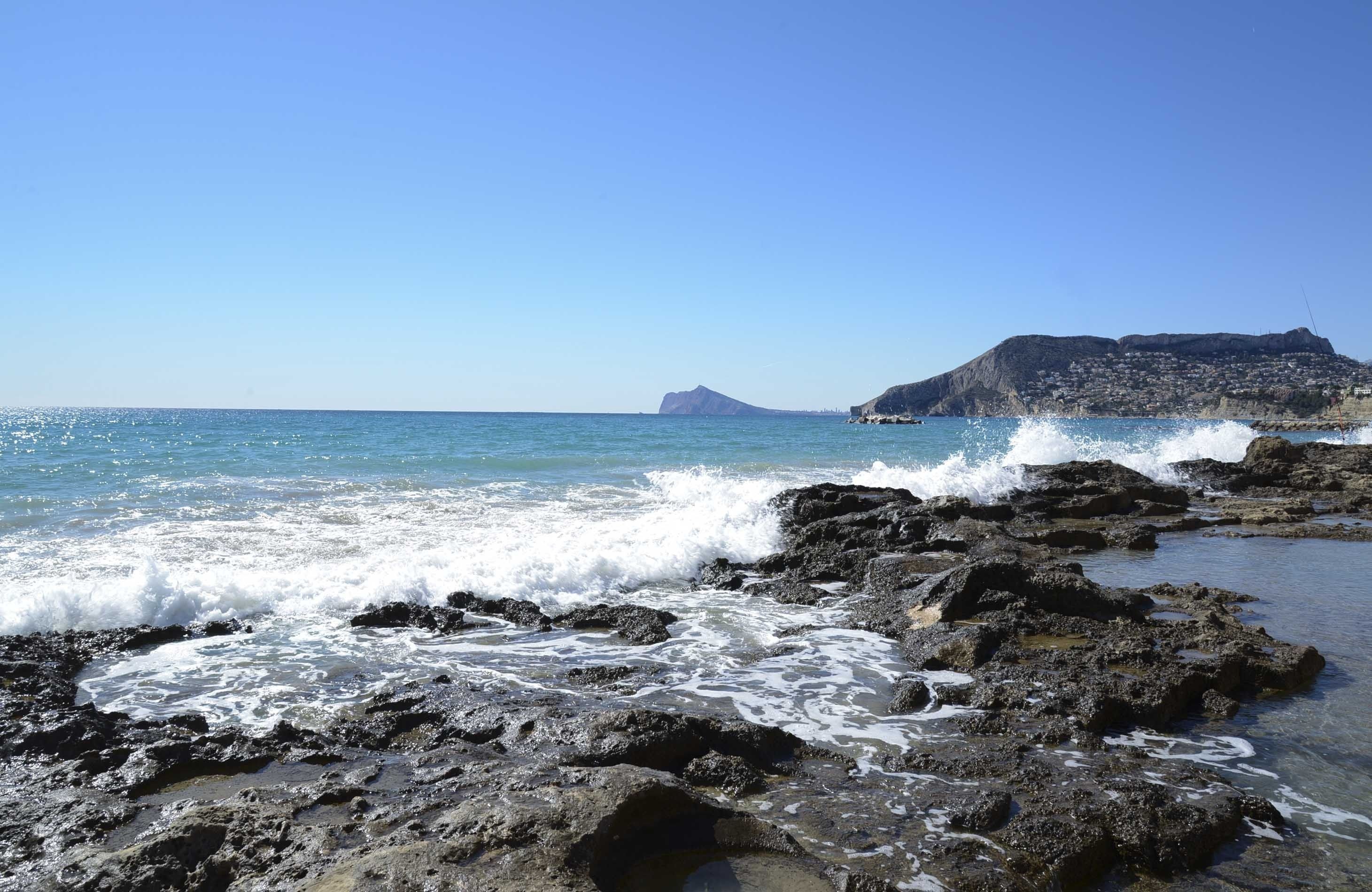 Una costa rocosa con olas rompiendo contra las rocas y una montaña distante con un pueblo en sus laderas.