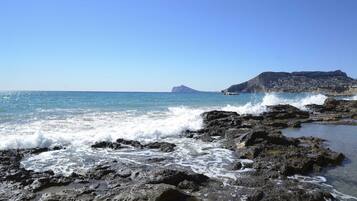 Una costa rocosa con olas rompiendo contra las rocas y una montaña distante con un pueblo en sus laderas.