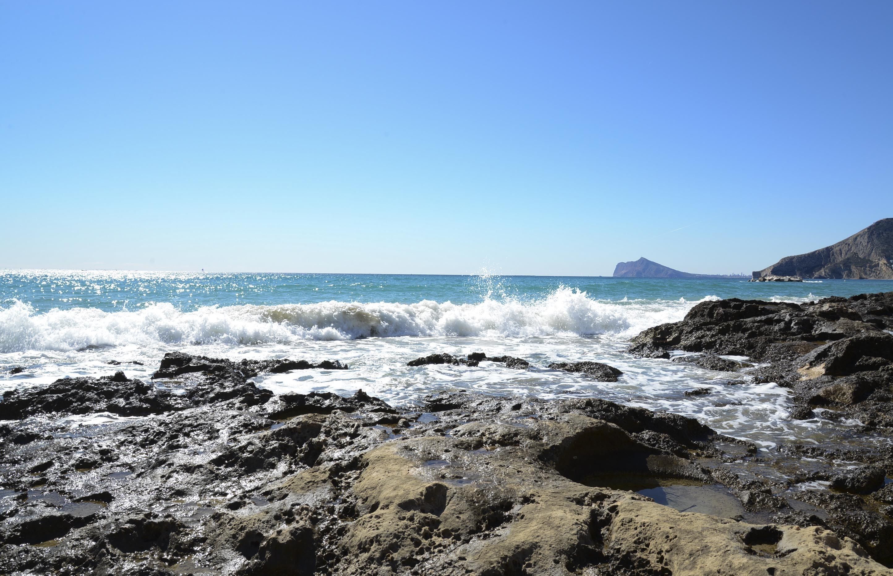 Un rivage rocheux où les vagues s’écrasent contre les rochers, sous un ciel bleu dégagé.