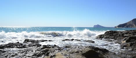 Un rivage rocheux où les vagues s’écrasent contre les rochers, sous un ciel bleu dégagé.