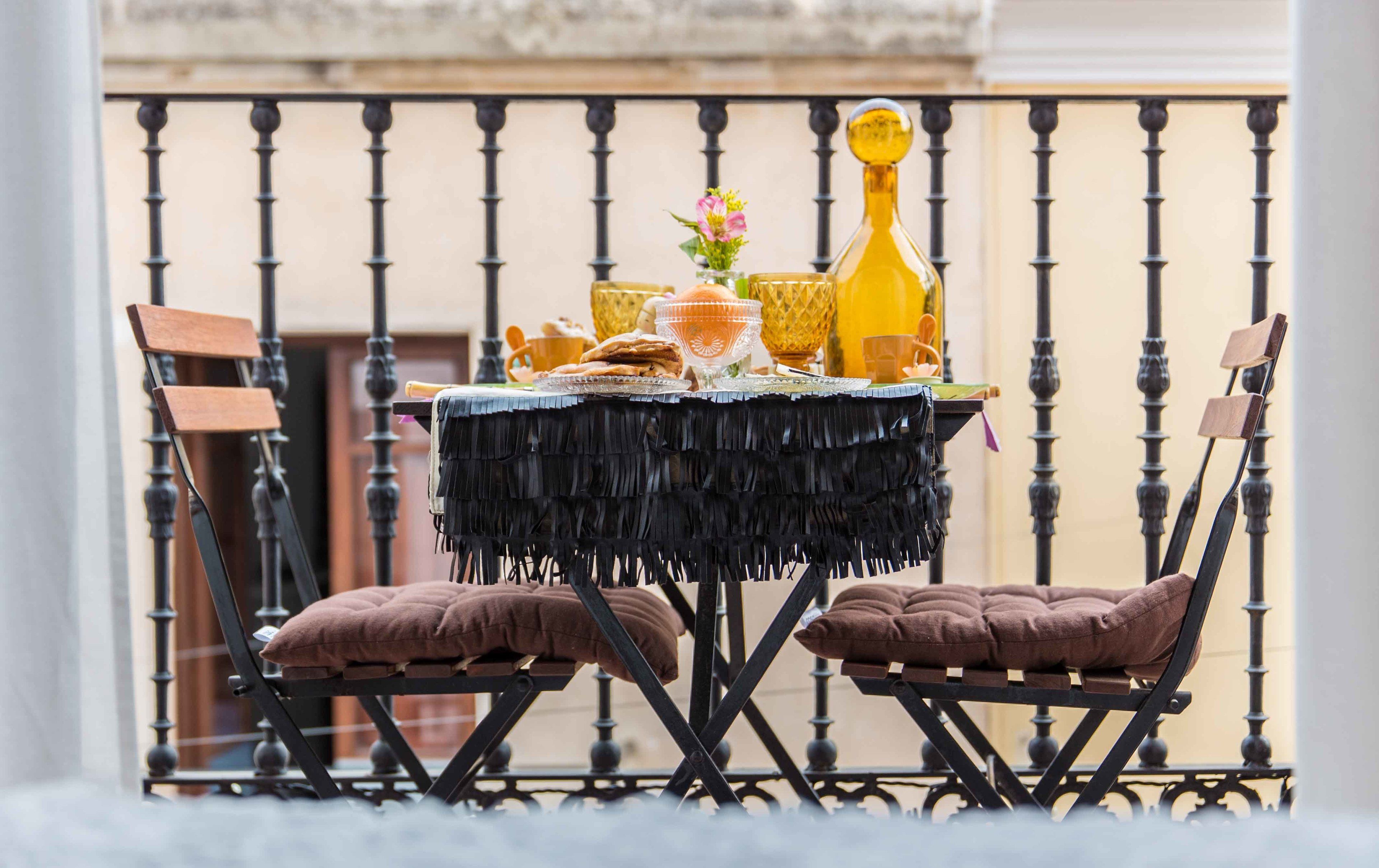A table set with a fringe tablecloth, two cushioned chairs, and various items including a yellow bottle, a glass, and a small vase with flowers.