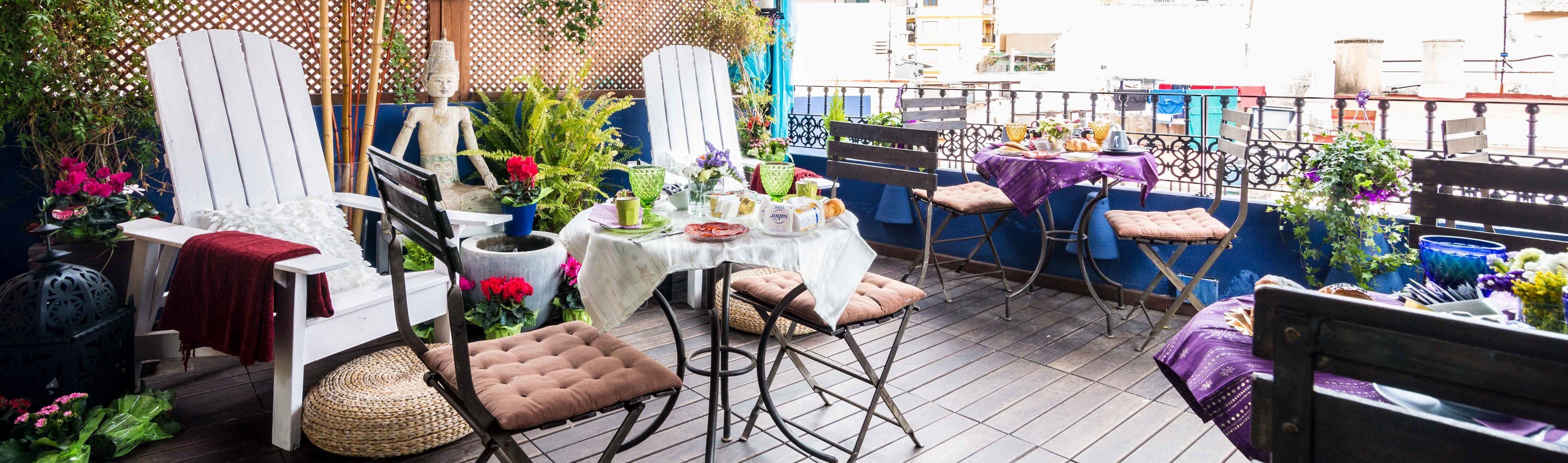 A patio with a table set for dining, surrounded by chairs and cushions, with plants and a decorative fence in the background.